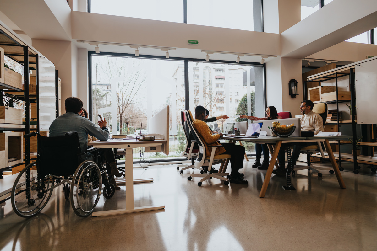 A multiracial group of employees collaborates around a table in a well-lit office space, showcasing wheelchair accessibility and teamwork.