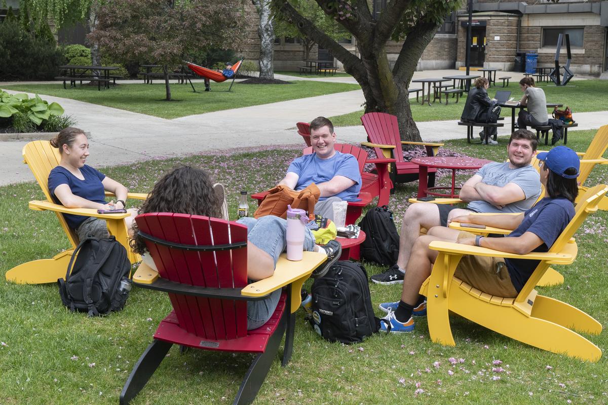 A group of five male and female students sit in Adirondack chairs on the quad