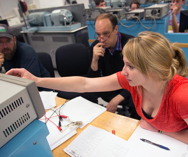 Two students work on electrical engineering equipment, supervised by a professor. 