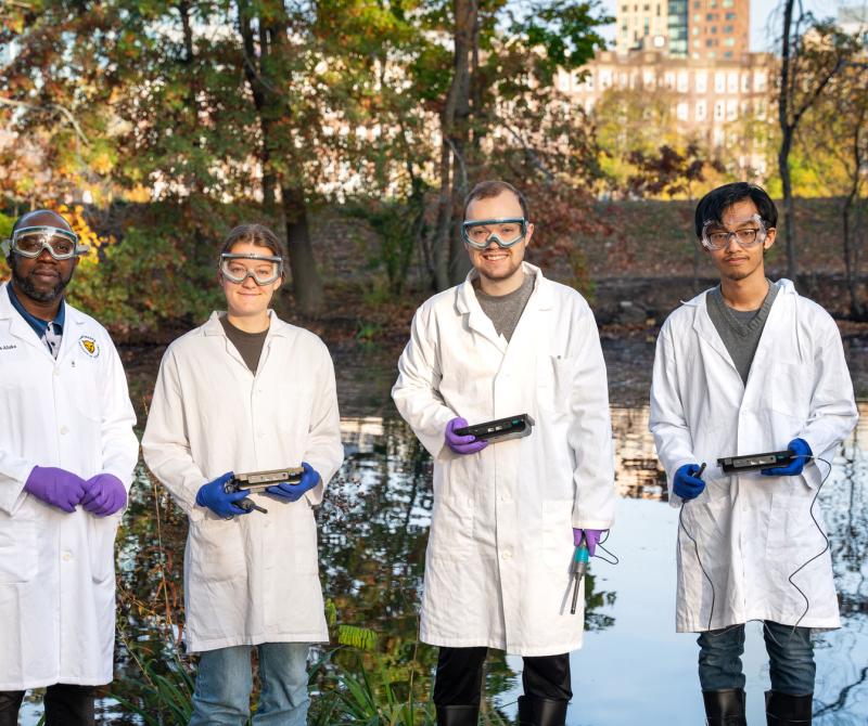 A faculty member and three students wearing lab coats stand by a body of water for measurements.