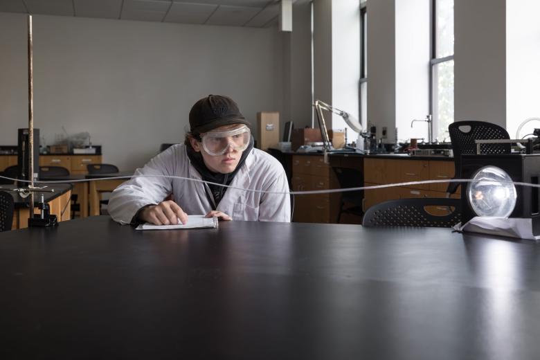 A male student wearing a lab coat and protective goggles conducts a period and frequency experiment in the Optics Lab