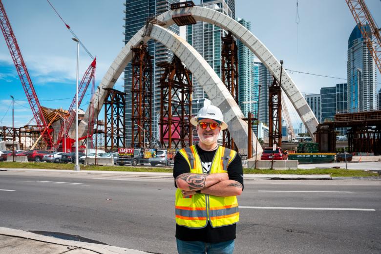 Jason Hatcher wearing a hardhat and safety vest stands in front a partially built bridge he is working on.