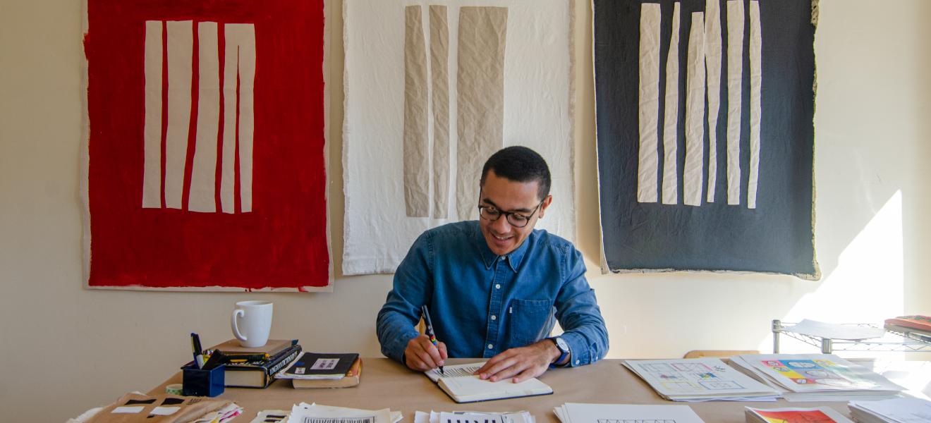 man drawing at a table with paintings in background